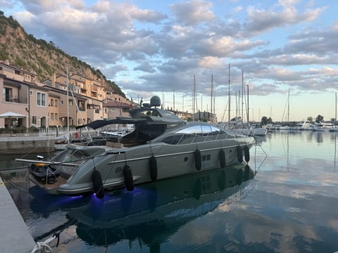 Luxury motor yacht docked in a Mediterranean marina with sailboats and waterfront buildings under cloudy sky
