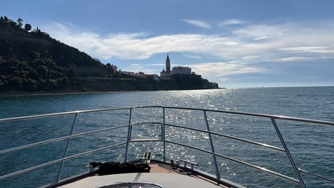 View from a boat deck with metal railings overlooking calm turquoise water toward a coastal town with a church steeple and green hillside